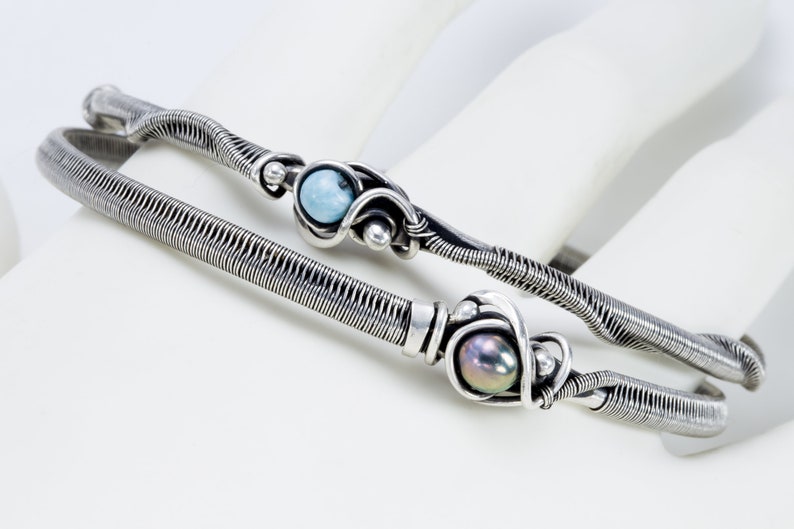 K&ouml;nnte beinhalten: Zwei silberne, drahtgewickelte Armb&auml;nder mit einem blauen Edelstein und einer schwarzen Perle. Die Armb&auml;nder liegen auf einer wei&szlig;en Oberfl&auml;che.