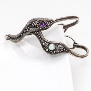 Könnte beinhalten: Ein silberfarbenes Armband mit einem einzigartigen, fließenden Design. Das Armband verfügt über einen Amethysten und einen Opal, jeweils in einer strukturierten, drahtumwickelten Fassung. Das Design beinhaltet kleine, perlenbesetzte Akzente.