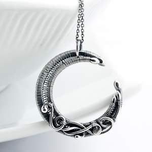 May include: A silver wire-wrapped crescent moon pendant necklace. The pendant is made of silver wire that is woven together to create a delicate and intricate design. The pendant is suspended from a silver chain.