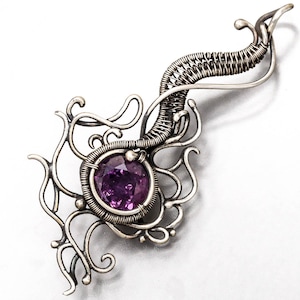 May include: A silver wire wrapped pendant with a purple gemstone in the center. The pendant is shaped like a stylized flower with intricate details.