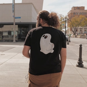 May include: A black t-shirt with a white ghost graphic on the back. The ghost has a smiling face and large eyes. The ghost is wearing a flower on its backside.