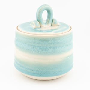 May include: A cylindrical ceramic jar with a lid featuring a prominent loop handle. The exterior displays horizontal bands of light blue glaze alternating with white, creating a textured, layered appearance on the surface of the container.