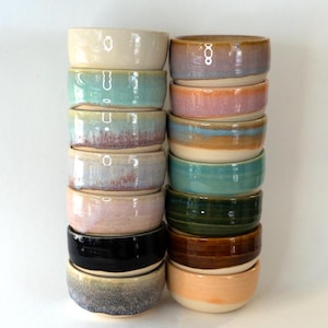 以下が含まれることがあります： クリーム、ティール、ピンク、黒、茶色など、さまざまな色の手作りのセラミックボウルのスタック。 ボウルはシンプルで丸みを帯びたデザインで、積み重ねられています。 ボウルは、食べ物を提供したり、装飾品として使用したりするためのものです。