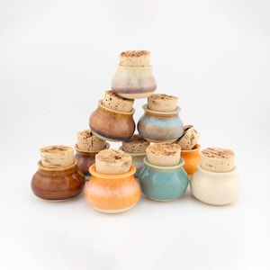 May include: A collection of small, round ceramic jars arranged in a pyramid stack. Each jar features a smooth, glazed finish in various colors including blue, orange, brown, and cream, and is topped with a cylindrical natural cork stopper.