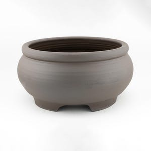 以下が含まれることがあります： 滑らかでマットな質感の、丸いダークブラウンの陶器製ボウル。幅広で曲線的なボディ、厚みのある丸みを帯びた縁、そして底面に小さな切り込みが入った凹型のベースが特徴で、安定感のある佇まいを見せています。