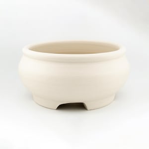Puede incluir: Un cuenco de cerámica de color blanco roto con un acabado liso y mate. El cuenco presenta una abertura ancha con un borde ligeramente curvado, un cuerpo redondeado y una base con pequeñas aberturas uniformes en el borde inferior.