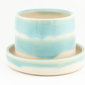 May include: A ceramic planter with a matching saucer, featuring a cylindrical shape and a glossy light blue glaze that drips over a cream-colored base. The glaze creates horizontal bands of color around the exterior of the pot and the rim of the saucer.