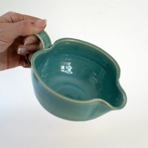 以下が含まれることがあります： 取っ手付きのターコイズブルーのセラミック製注ぎ口付きボウル。ボウルは滑らかで光沢のある仕上げで、わずかに丸みを帯びた形をしています。