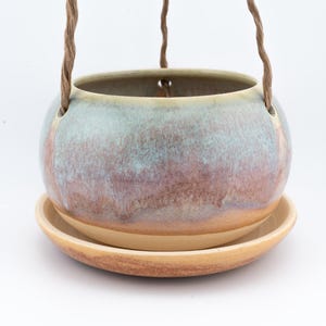 May include: A ceramic hanging planter with a round, speckled blue and purple body and a matching saucer. The planter is suspended by twisted brown rope. The planter is designed for indoor or outdoor use.