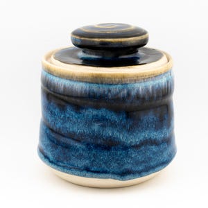 May include: A ceramic lidded jar featuring a deep blue and black glaze with a glossy finish. The cylindrical body transitions to a lighter cream-colored base, while the matching lid has a central knob and a dark, swirling glaze pattern.