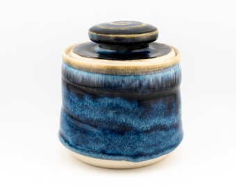 May include: A ceramic lidded jar featuring a deep blue and black glaze with a glossy finish. The cylindrical body transitions to a lighter cream-coloured base, while the matching lid has a central knob and a dark, swirling glaze pattern.