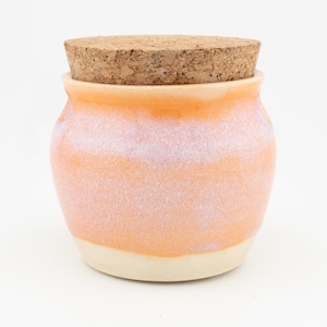 Op de afbeelding: Een kleine keramische pot met een afgerond lichaam, voorzien van een kleurverloopglazuur dat overgaat van zacht perzik-oranje aan de bovenkant naar een romige gebroken witte basis. De pot is afgesloten met een rond, getextureerd deksel van natuurkurk.