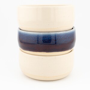 May include: A stack of three ceramic bowls. The top and bottom bowls are a solid off-white color with a smooth finish. The middle bowl features a deep blue glaze that transitions into a darker navy tone, creating a distinct contrast in the stack.
