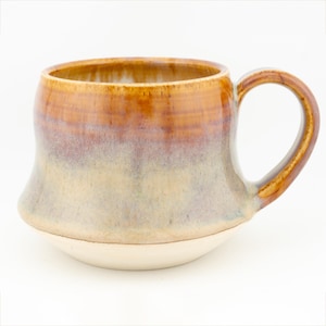 May include: A handmade ceramic mug with a unique shape and a large handle. The mug features a gradient of colours, transitioning from a light cream base to a light purple, then to a warm brown at the rim. The mug is approximately 10 cm tall.