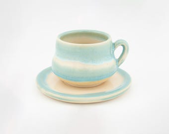 May include: A ceramic cup and saucer set featuring a glossy glaze with a gradient of light blue and cream white. The cup has a rounded body and a curved handle, while the matching saucer has a smooth, circular rim with a blue and cream finish.