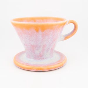 May include: A ceramic coffee dripper featuring a conical shape with a wide rim and a single handle. The exterior is finished with a reactive glaze in shades of soft pink, white, and vibrant orange, creating a marbled effect across the surface.