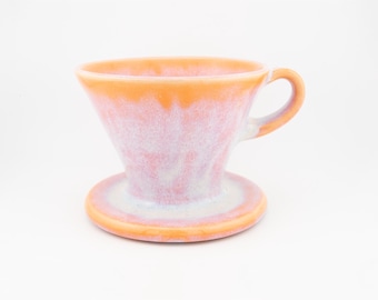 May include: A ceramic coffee dripper featuring a conical shape with a wide rim and a single handle. The exterior is finished with a reactive glaze in shades of soft pink, white, and vibrant orange, creating a marbled effect across the surface.