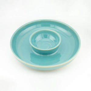 Op de afbeelding: Een ronde, turquoise keramische serveerschaal met een brede, platte buitenrand en een kleinere, diepe ronde kom in het midden. Het object heeft een gladde, glanzende afwerking met een lichtgekleurde rand langs de buitenkant en de kom.
