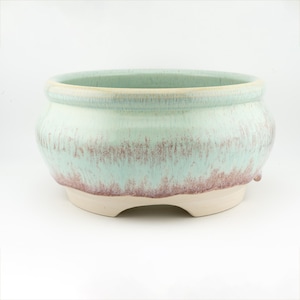 May include: A round ceramic plant pot with a pale green glaze and reddish-brown accents. The pot has a wide opening, a rounded body, and three small feet. The glaze has a textured finish, suitable for indoor or outdoor use. Measurements are in centimeters.