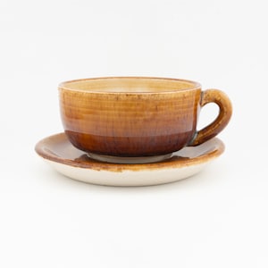 May include: A ceramic cup and saucer set featuring a glossy, variegated brown glaze that transitions from a deep, dark brown at the base to a lighter, honey-toned brown at the rim. The saucer has a matching brown rim with a cream-colored center.