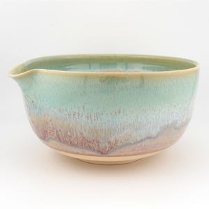May include: A handmade ceramic mixing bowl with a pouring spout. The bowl features a light green glaze that fades into a reddish-brown and cream color. The bowl is ideal for kitchen use.