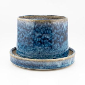 May include: A cylindrical ceramic planter with a deep blue and mottled navy glaze, featuring a smooth rim and a matching circular saucer base. The finish displays a variegated texture with lighter blue and beige tones along the edges.