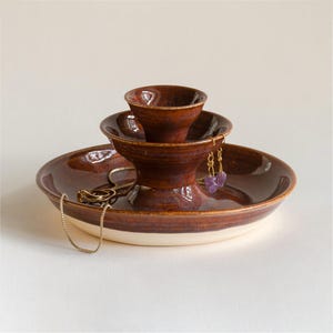 Peut inclure: Un plat à bijoux en céramique à deux niveaux avec un vernis brun tacheté brillant et une base crème. La structure comprend un large plateau inférieur et un bol surélevé plus petit. Une chaîne dorée et des boucles d'oreilles violettes reposent dessus.