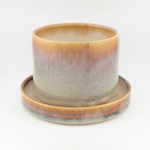 Pode incluir: Um vaso cilíndrico de cerâmica com um pires circular a condizer. A peça apresenta um esmalte reativo com um gradiente que transita de um castanho-mel quente no rebordo para tons malva e cinzentos suaves na base, com acabamento liso.