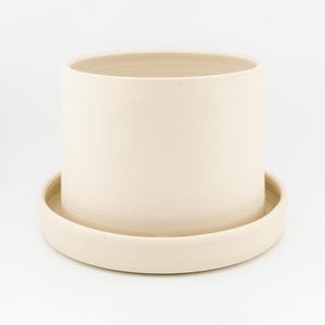 Puede incluir: Una maceta cilíndrica de cerámica en color blanco roto con un acabado mate suave, que incluye una base de plato circular a juego. El recipiente tiene paredes verticales rectas y una parte superior abierta, ofreciendo una estructura simple.