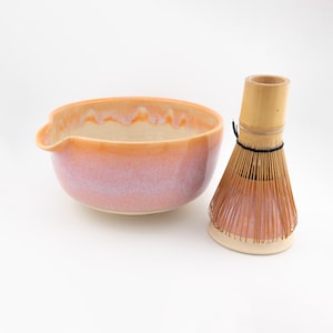 Può includere: Una ciotola in ceramica per matcha con smalto sfumato rosa e arancione e un beccuccio, abbinata a un frustino da tè in bambù con setole sottili color marrone chiaro legate da un filo nero, su uno sfondo bianco uniforme.