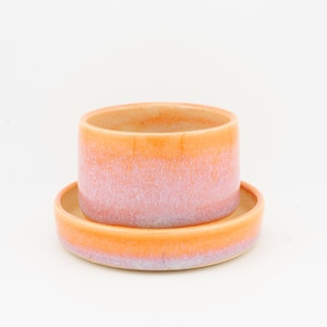 May include: A ceramic planter with a matching saucer. The planter and saucer are cylindrical and feature a gradient of colours, transitioning from orange to pink and lavender. The design is simple and modern.