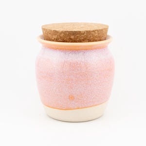 Op de afbeelding: Een kleine keramische pot met een rond lichaam en een gespikkeld perzik en wit glazuur. De pot heeft een brede opening aan de bovenkant en is afgesloten met een rond, lichtbruin kurken deksel. De onderkant is effen gebroken wit van kleur.