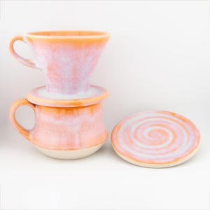 Può includere: Un set per versare il caffè in ceramica nei toni dell'arancione e del bianco. Il set comprende un versatore a forma di cono, una tazza con manico e un piccolo sottobicchiere rotondo con un motivo a spirale. I pezzi hanno una finitura macchiettata e smaltata.