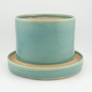 May include: A cylindrical ceramic planter with a smooth, glossy teal glaze on the exterior and a matching circular saucer base. The interior of the pot and the top surface of the saucer reveal a natural, unglazed light beige clay finish.