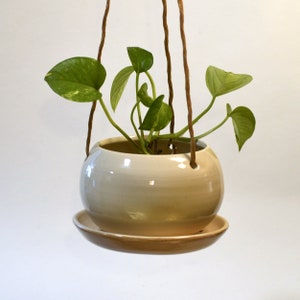 Puede incluir: Un macetero colgante con una maceta de cerámica beige claro y un platillo a juego. La maceta es redonda y tiene un acabado liso y brillante. Una planta verde con enredaderas colgantes crece en la maceta.