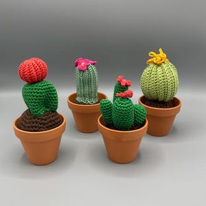 Op de afbeelding: Vier gehaakte cactussen in terracotta-kleurige potten. Eén heeft een rode top, één een roze bloem, één heeft rode bloemen en één heeft een gele bloem. Elke cactus staat in bruine gehaakte aarde in de kleine, ronde, oranje-getinte potten.