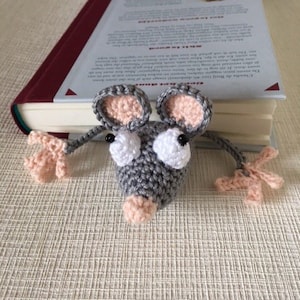 Op de afbeelding: Een grijze gehaakte muis boekenlegger met roze oren en neus. De muis houdt een boek vast met een witte kaft.