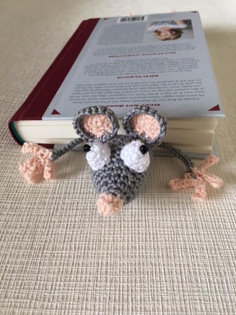 Op de afbeelding: Een grijze gehaakte muis boekenlegger met roze oren en neus. De muis houdt een boek vast met een witte kaft.