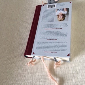 Op de afbeelding: Een grijze en roze gehaakte muis bladwijzer met lange roze garenstaarten. De bladwijzer is geplaatst op de rug van een boek met een witte omslag en zwarte tekst.
