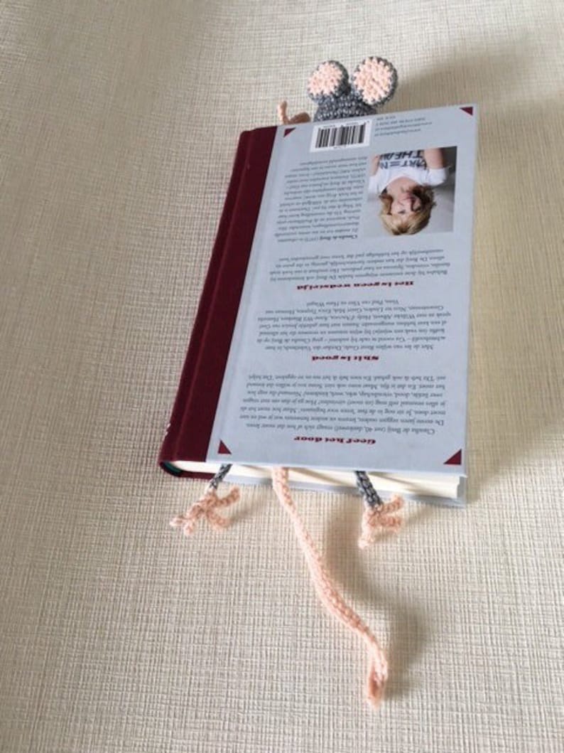 Op de afbeelding: Een grijze en roze gehaakte muis bladwijzer met lange roze garenstaarten. De bladwijzer is geplaatst op de rug van een boek met een witte omslag en zwarte tekst.