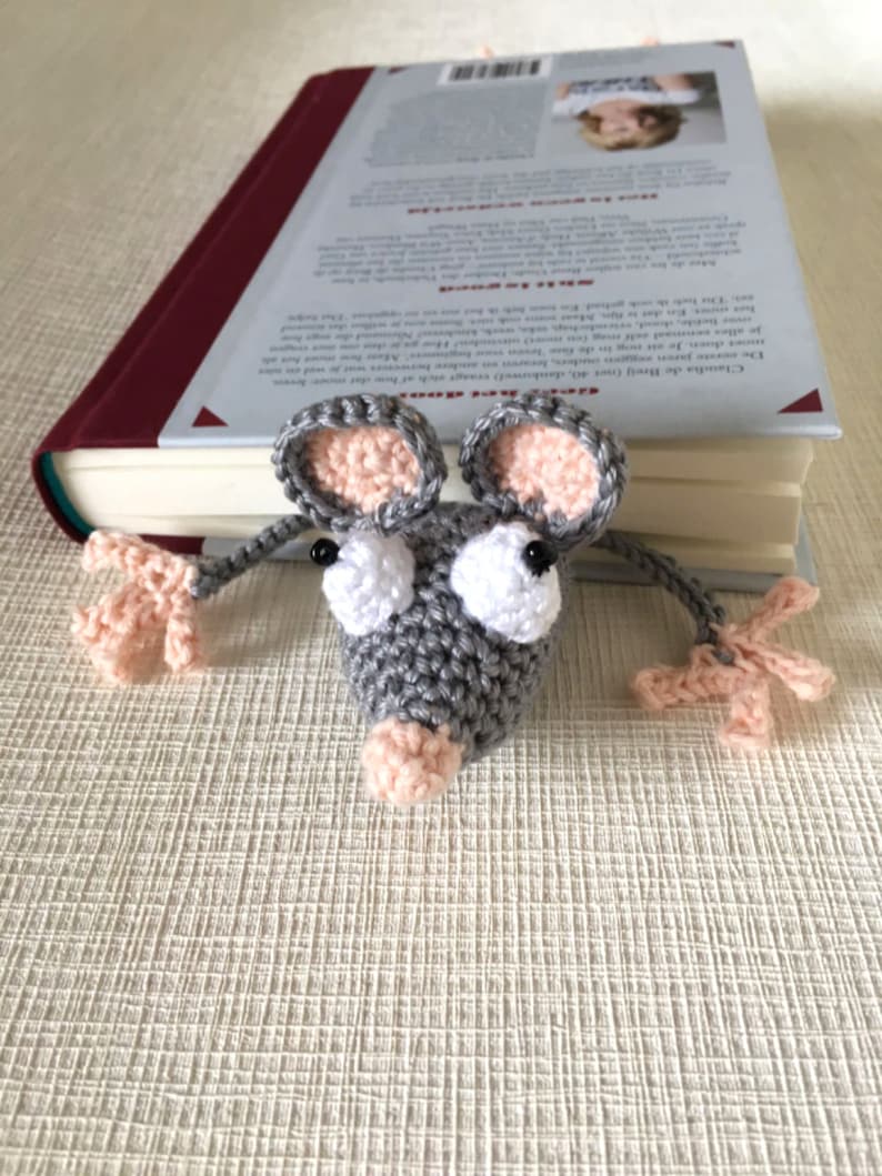 Op de afbeelding: Een grijze gehaakte muis boekenlegger met roze oren, neus en voeten. De muis heeft grote witte ogen en zit bovenop een boek.