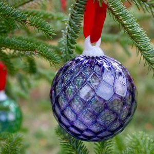 May include: A purple glass Christmas ornament with a textured, diamond-like pattern hangs from a green pine tree branch. The ornament is decorated with iridescent accents.