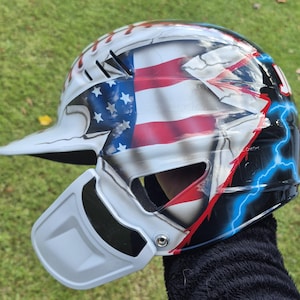 May include: A baseball catcher's helmet with a patriotic design. The helmet features the American flag, lightning bolts, and a white and gray color scheme. The helmet has a visor and a face guard.