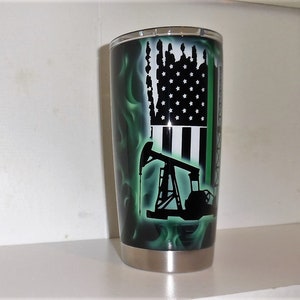 May include: A stainless steel tumbler with a green and black design. The design features an American flag, an oil derrick, and a thin green line.