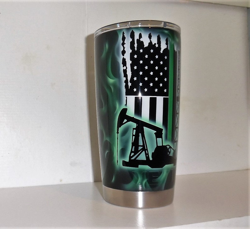 May include: A stainless steel tumbler with a green and black design. The design features an American flag, an oil derrick, and a thin green line.