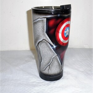 May include: A stainless steel tumbler with a black lid and a design featuring Captain America's shield on a cracked, gray background.