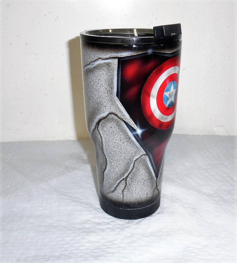May include: A stainless steel tumbler with a black lid and a design featuring Captain America's shield on a cracked, gray background.