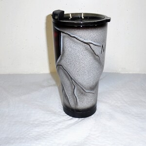 May include: A white and black stainless steel tumbler with a black lid and a white and black marble design. The tumbler has a black base and a white lightning bolt design.