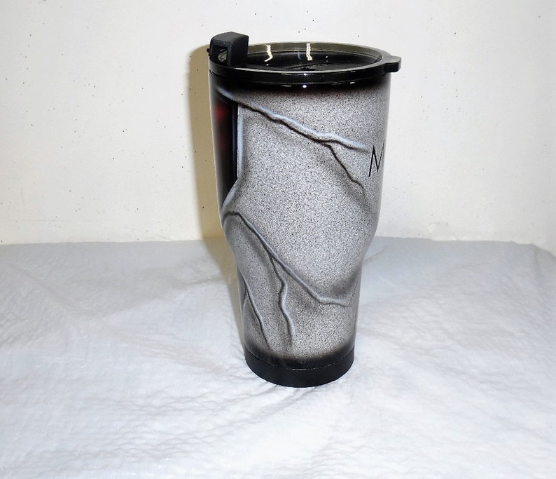 May include: A white and black stainless steel tumbler with a black lid and a white and black marble design. The tumbler has a black base and a white lightning bolt design.