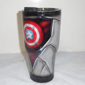 May include: A stainless steel tumbler with a black lid and a graphic of Captain America's shield on a cracked, gray background.